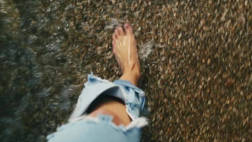 A woman wearing torn blue jeans is walking barefoot along the coast as waves roll in over her feet