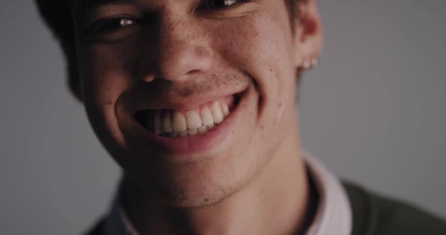 Handsome young man wearing earrings smiling and standing in front of a white wall at a studio