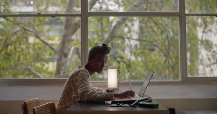 A woman is sitting at a desk working on a laptop computer and writing on a tablet