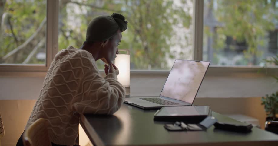Black woman wearing a head wrap sitting at the desk next to the window using her laptop during daytime