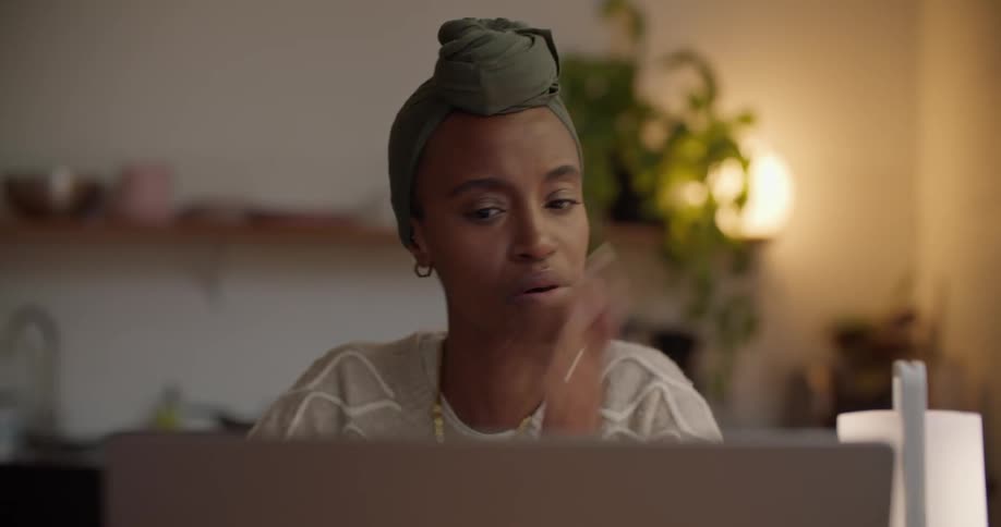 Thoughtful black woman wearing a head wrap sitting at the desk and using her laptop indoors