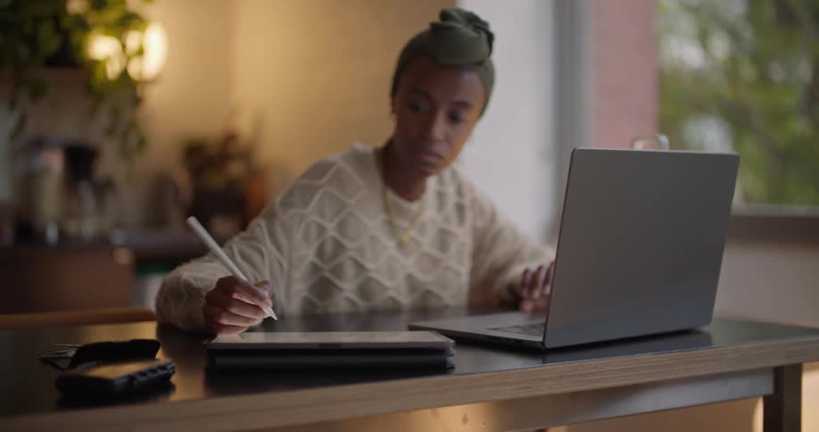 Black woman wearing a head wrap sitting at the desk near the window writing with a pen while using her laptop