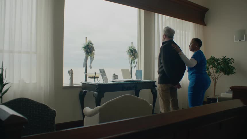 A man standing before a window while gazing outside is approached by a nurse who takes his hand and begins to speak to him