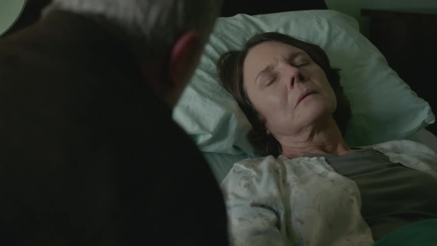 Old man sitting next to his dying wife and talking to her while she is asleep on the bed