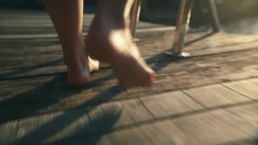 Person walks barefoot across wooden deck and turns to face water near metal ladder