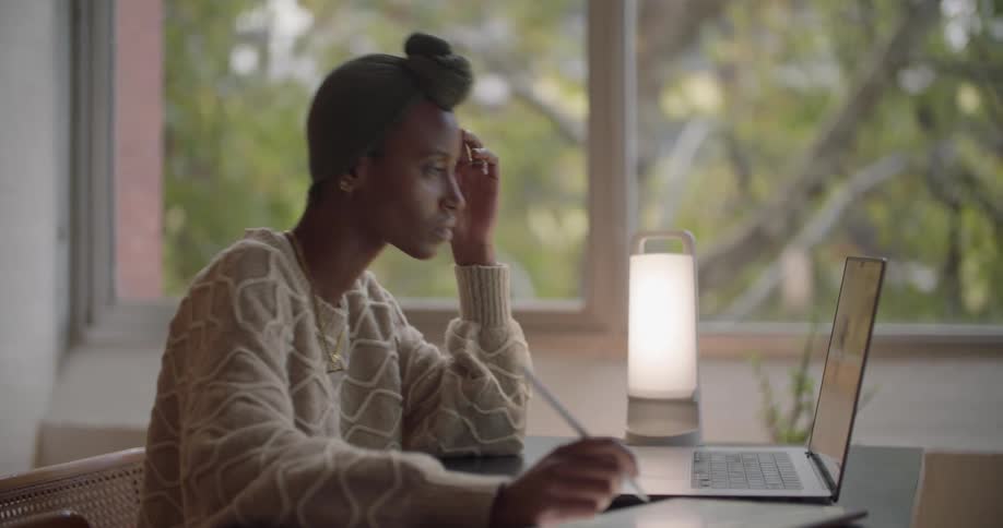 Black woman wearing a head wrap sitting at the desk next to the windows using her laptop and writing with a pen
