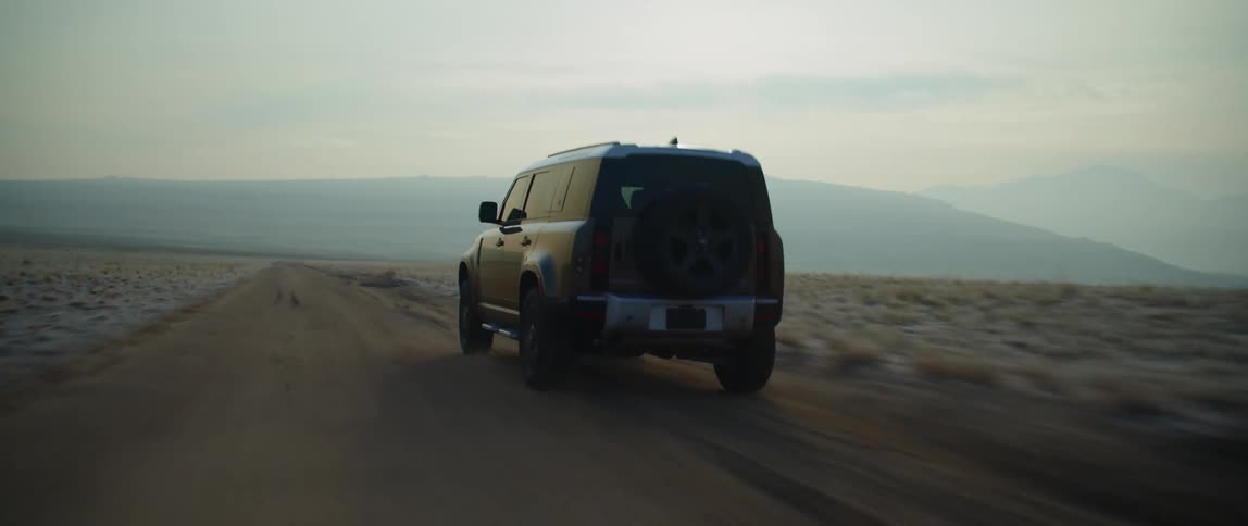 A black truck is traveling on a dirt road and there are mountains in the distance under a hazy sky
