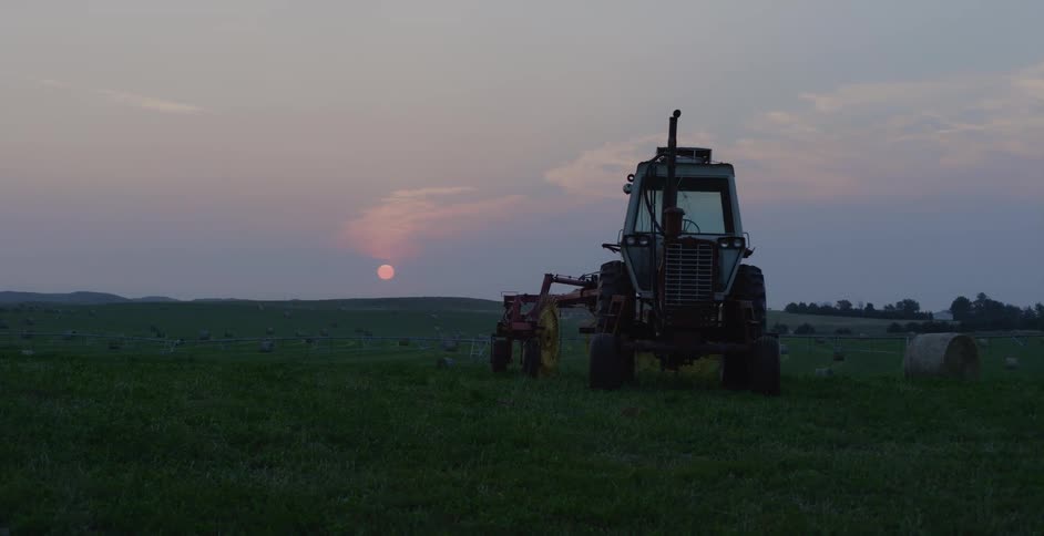 A tractor sits idle in a field with mountains in the distance as the sun sets