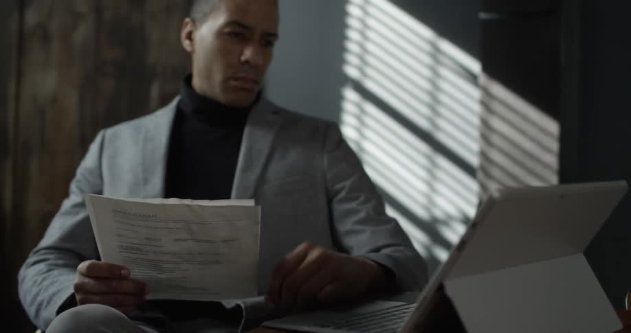 A man reading  some papers and typing on a laptop