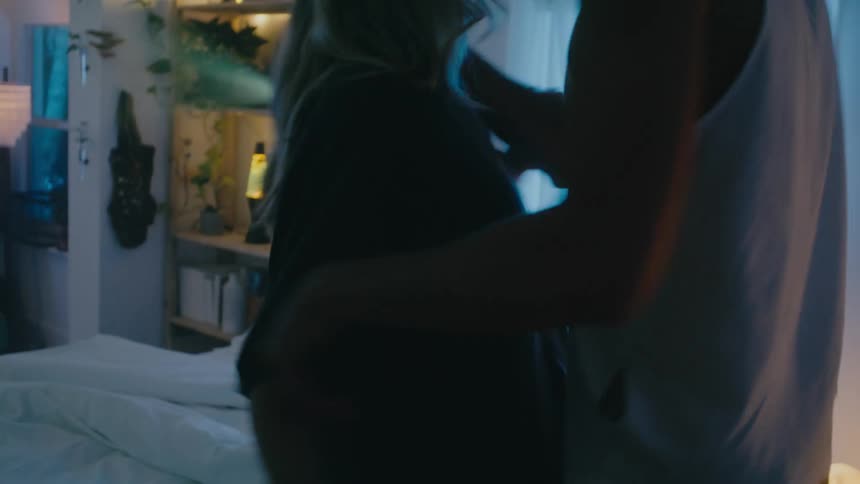 A woman leads a man into a bedroom and pushes him onto the bed before the two begin to kiss