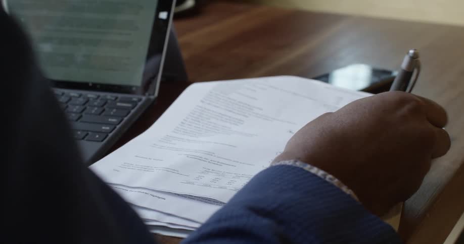 A woman is working at a laptop and taking notes on some paperwork