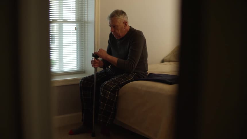 An old man sits on the edge of a bed and grasps his walking cane