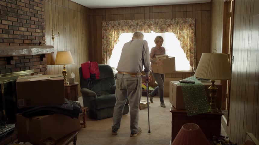 A man is leaning on a cane as a woman carries boxes out