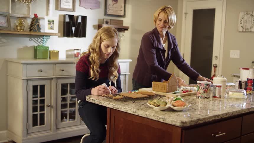 A girl is preparing food at a table as a woman puts a tray next to her smiling