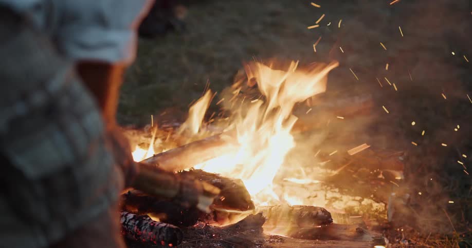 A person is standing by a bonfire and putting logs in it