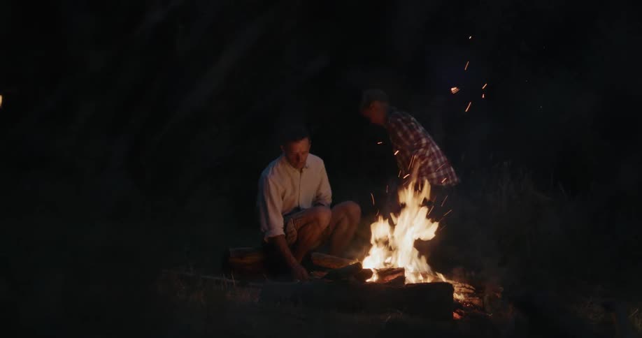 A boy sticks wood in a campfire then sits down and the dad pokes it with a long stick