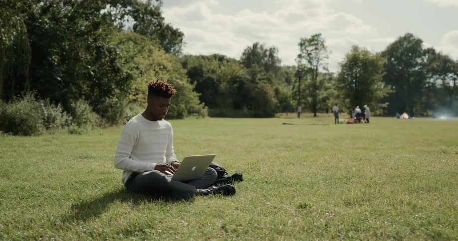 A man sits in the park on the grass typing on his computer while a small crowd interacts in the distance