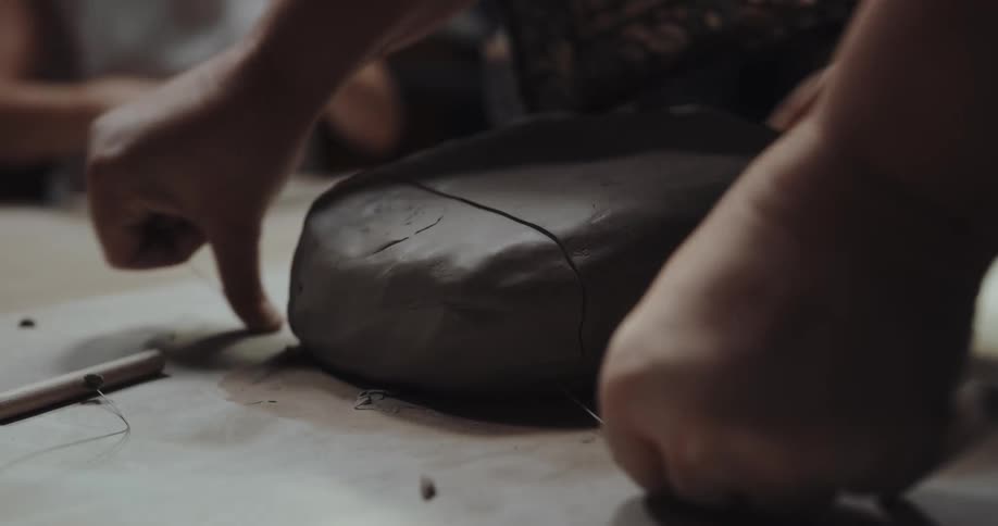 Hands use a thread to slice a block of clay while a teenager pats and softens a brick of clay
