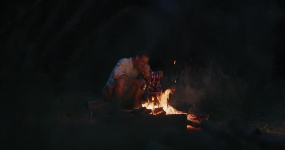 A dad hugs his son as they sit around the campfire at night