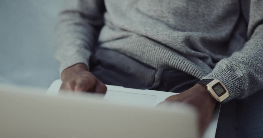 Black man picking up his laptop from the couch sitting down and using it on his lap