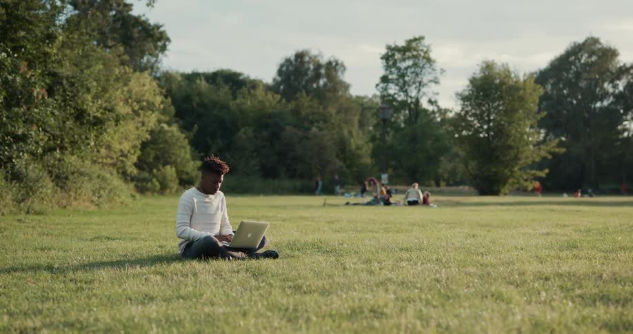 A man is sitting on the grass typing on a laptop computer and there are people sitting in the distance