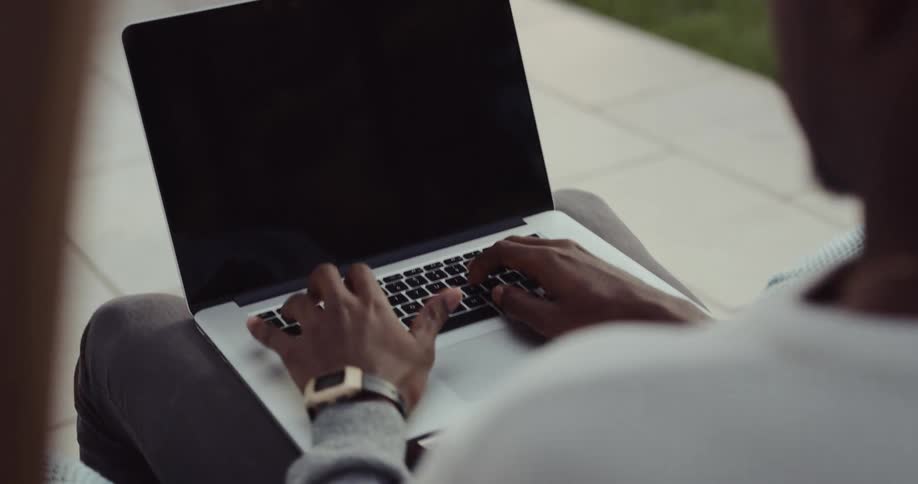 A man is typing on a laptop and sitting outdoors