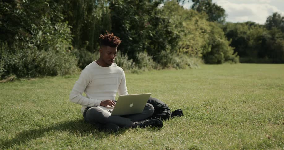 A man is sitting outside on the grass typing on his laptop computer