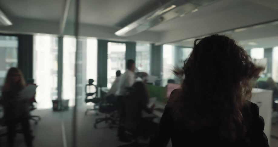 A woman checks her watch while walking quickly through an office towards her desk