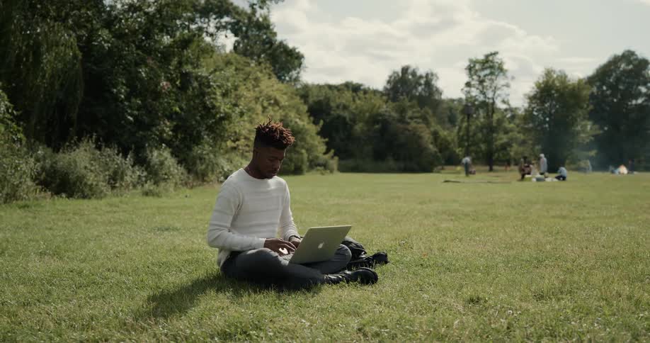 A man is sitting on the grass typing on a laptop computer and there are people in the distance