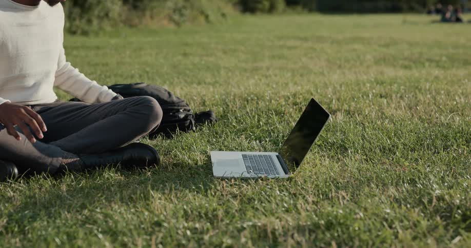 A man is sitting outside in the grass with his laptop computer and he sets it on the grass and picks up his cell phone