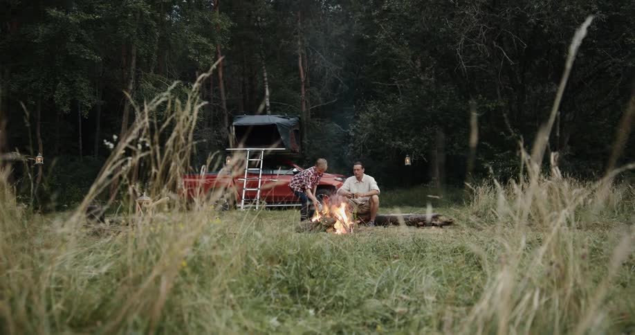 Two males sit around a campfire in a woodland area