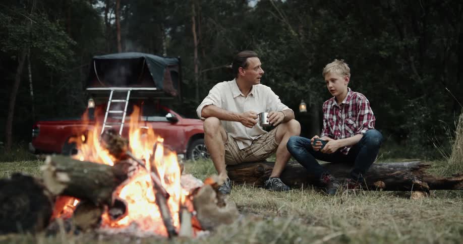 Father with white shirt and brown shorts sits on log with son next to campfire