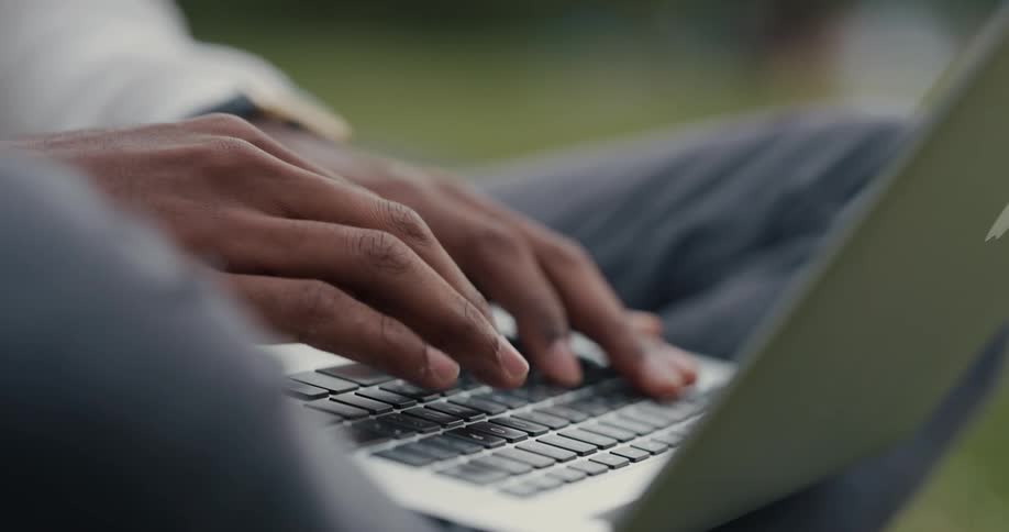 A man is sitting on the grass typing on a laptop computer that is sitting on his legs