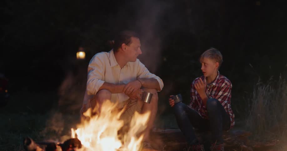 Father and son are sitting by a bonfire outdoors at night