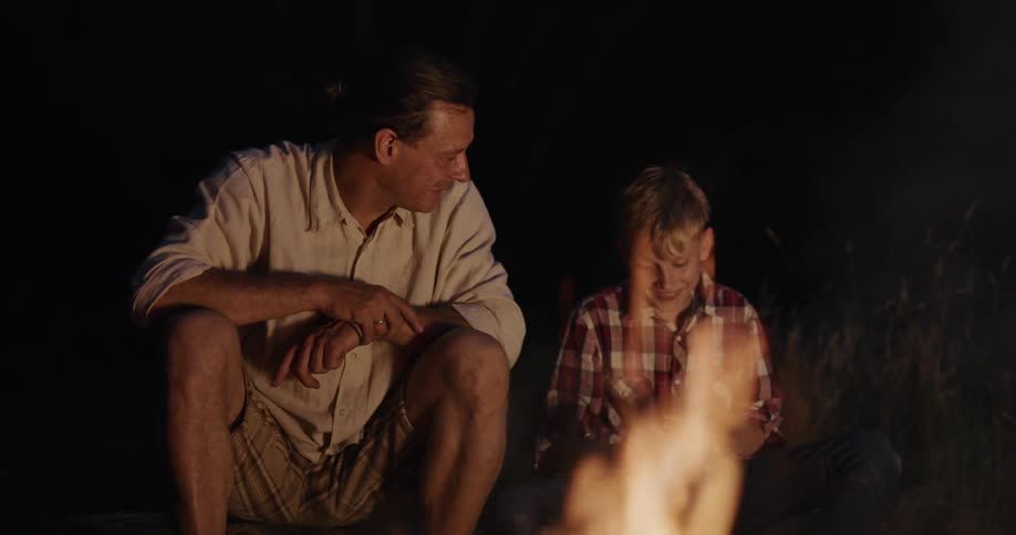 A dad throws another log onto the fire then continues to talk and laugh with his son