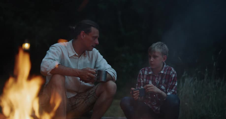 Father with white shirt and brown shorts sits next to son by campfire at night