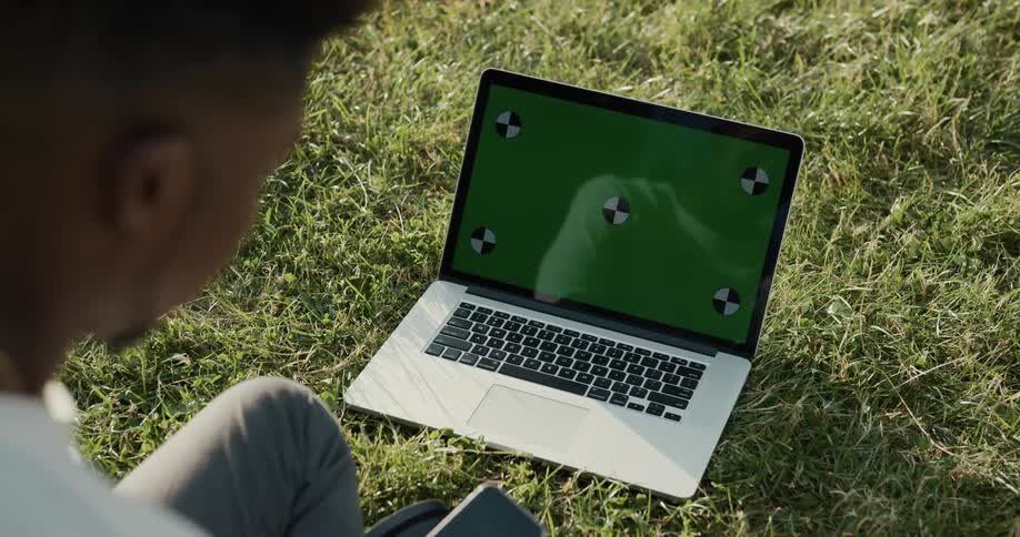Man with gray jeans and white shirt looks down at laptop screen on green lawn