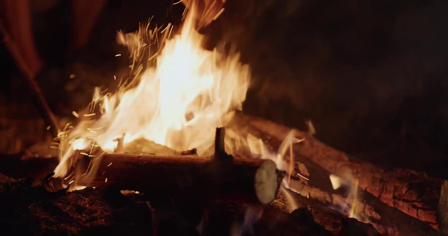 A man stokes a campfire causing embers and sparks to fly up from the flames