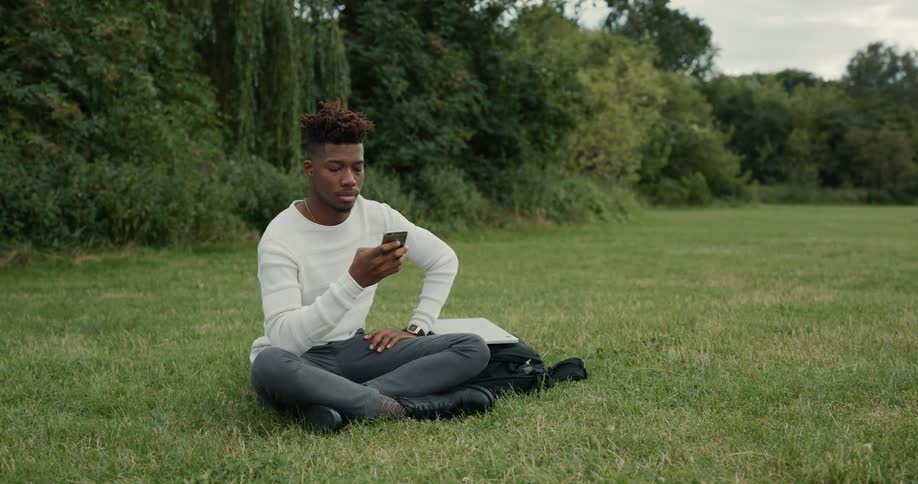 A man is sitting outside in a field near woods holding his cell phone
