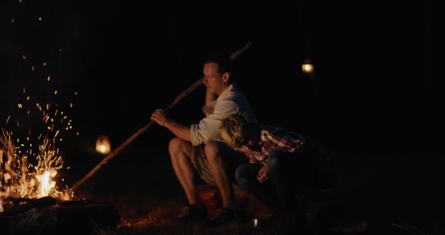 A boy walks up to a campfire and puts a log in as the dad sits next to it
