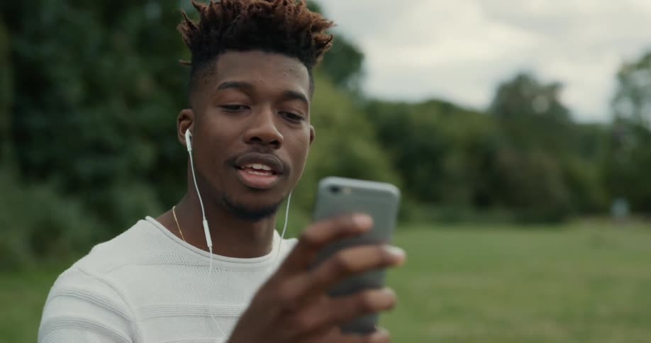 Man standing outdoors in field bordering woods puts on earbuds and has a video call on his smartphone