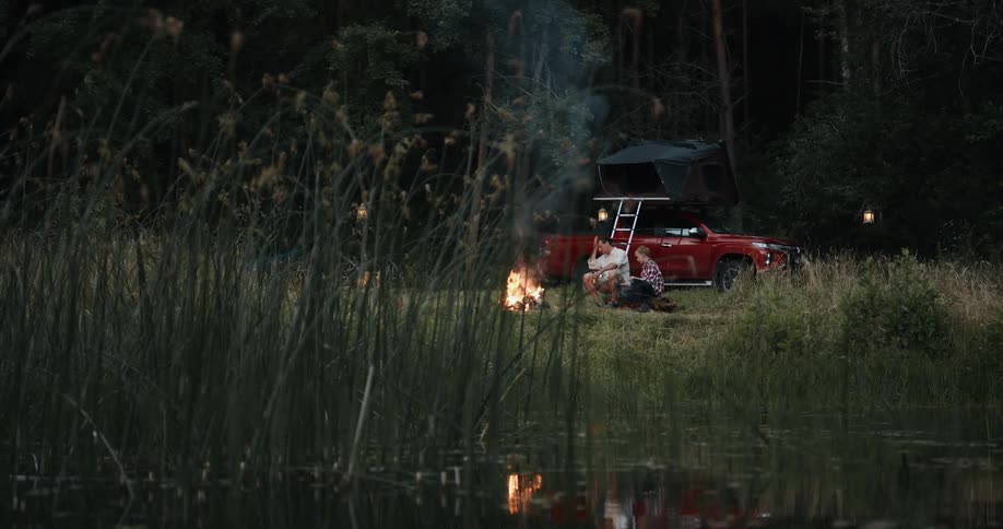 A father and son tend a campfire together on the shore of a lake during the day