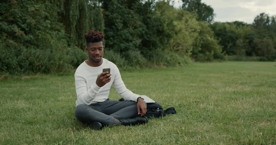 A man is sitting outside in a field near woods holding his cell phone