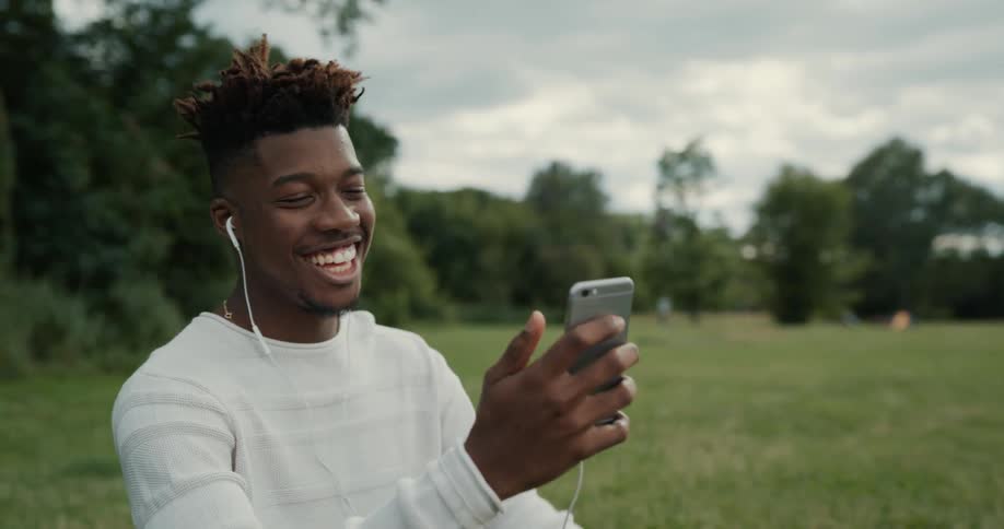 A man is sitting outside in a field near woods talking on his cell phone