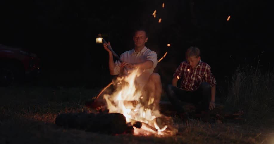 Father and son sit in front of a campfire while camping at night