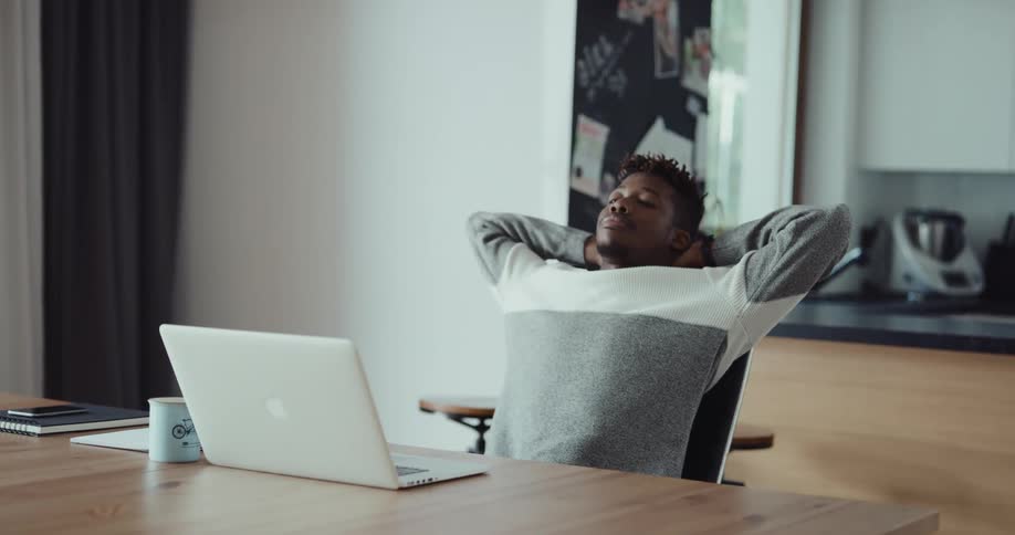 A man sits at his dining room table with his laptop in front of him as he takes a sip of coffee then stretches