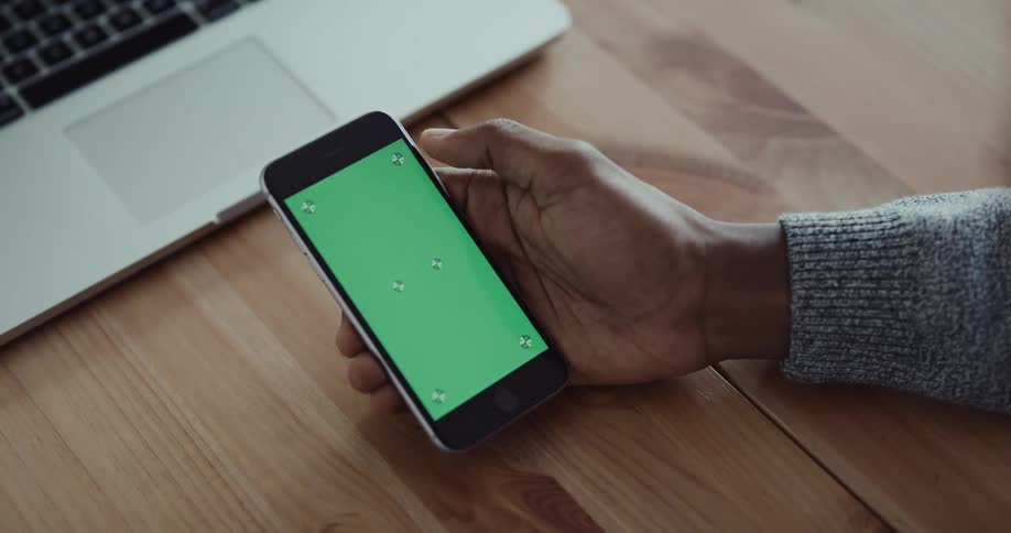 Black mans hand tapping on a smartphone screen he is holding in front of a wooden desk and a laptop