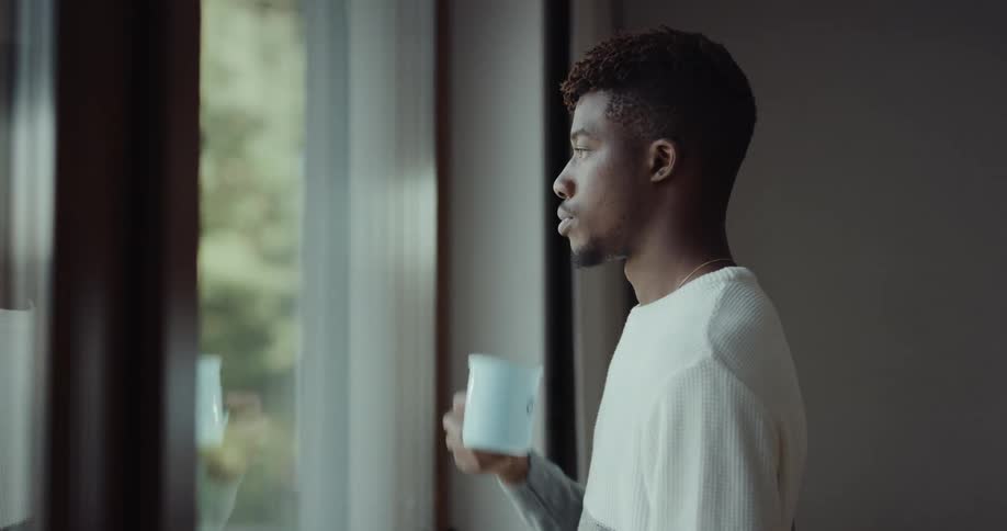 Contemplative young black man standing in front of the window drinking a cup of coffee