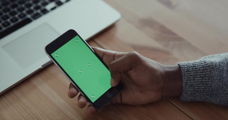 Black mans hands tapping on a smartphone screen in front of a wooden desk with a laptop on it