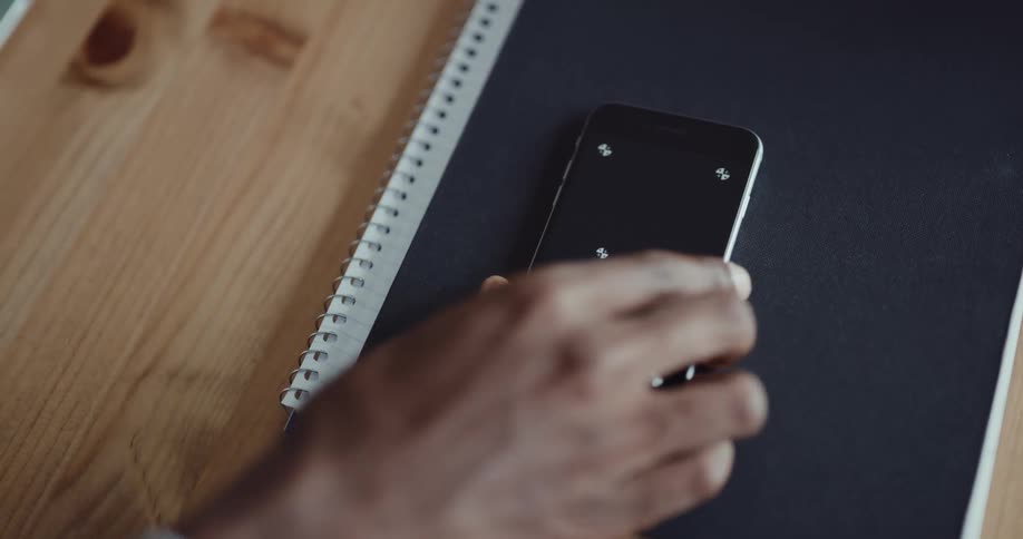 Black mans hand picking up his smartphone that was on top of a notebook on a wooden desk